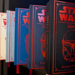 May include: A row of Star Wars books with red and black covers. The books have titles like "501st" and feature character illustrations. The spines display the author's name, Karen Traviss. Each book has a red bookmark.