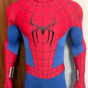 May include: A full-body spandex suit featuring a red and blue color scheme with a black spider emblem on the chest. The red sections have a black web pattern, while the blue sections are solid. Silver wrist-mounted accessories are attached.