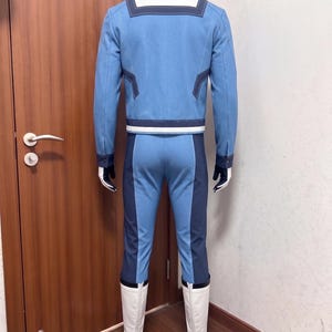 May include: A light blue long-sleeved jacket with a white collar and dark blue trim, paired with matching light blue trousers featuring dark blue side panels. The outfit includes white boots and dark gloves, presented on a mannequin from the back.