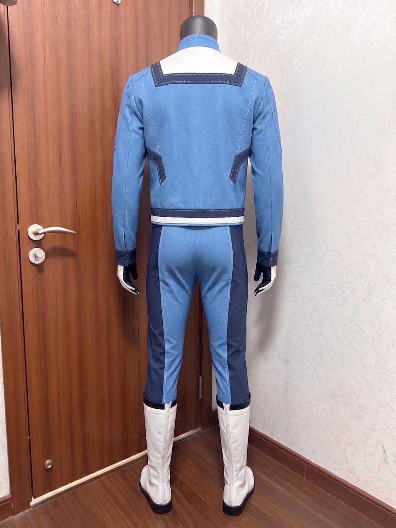 May include: A light blue long-sleeved jacket with a white collar and dark blue trim, paired with matching light blue trousers featuring dark blue side panels. The outfit includes white boots and dark gloves, presented on a mannequin from the back.