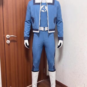 May include: A blue and white costume featuring a jacket with a number 4 emblem on the chest, matching blue trousers, a white belt, and white boots. The jacket has a high collar, dark blue accents, and a full-length zipper. The ensemble is displayed on a mannequin.