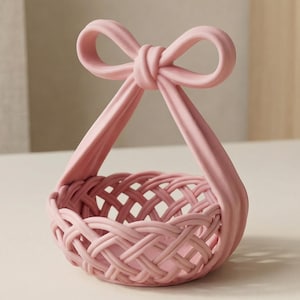 Puede incluir: Una pequeña cesta rosa con un diseño tejido y un asa en forma de lazo. La cesta está hecha de un material sólido y se encuentra sobre una superficie de color claro. El asa está unida a la cesta con dos soportes curvos.