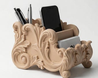 May include: A beige desk tidy with an ornate, scrollwork design. It holds pens, a smartphone, and business cards. The organiser has a classic, decorative style, ideal for a home office or desk.
