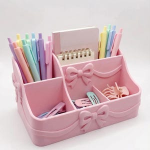 Könnte beinhalten: Ein rosa Schreibtisch-Organizer mit mehreren Fächern, verziert mit Schleifen. Der Organizer enthält Stifte, Haarspangen und einen kleinen Notizblock. Pastellfarbene Stifte und Clips sind sichtbar und schaffen einen koordinierten Look.
