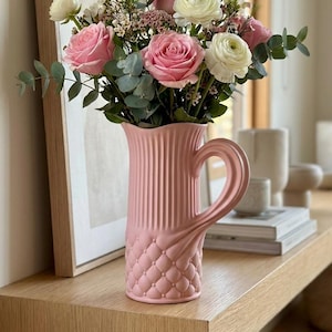 May include: A pink ceramic jug with a handle, holding a floral arrangement of pink roses, white flowers, and foliage. The jug features a quilted base and a ribbed upper section. The display is on a wooden shelf.
