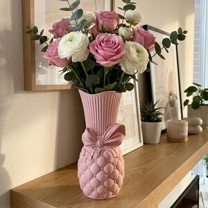 May include: A pink vase, decorated with a bow, displays a floral arrangement of pink roses, white ranunculus, and eucalyptus. The vase has a quilted pattern. The display sits on a wooden shelf, with framed art in the background.