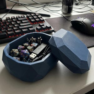 Puede incluir: Una caja de almacenamiento azul de forma geométrica, abierta para revelar dados, figuritas y una unidad USB. La caja está sobre un escritorio con un teclado, un ratón y un monitor de ordenador en el fondo. La caja mide aproximadamente 15 cm de largo.