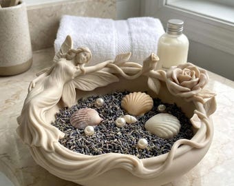 May include: A cream-coloured decorative bowl with a sculpted fairy design, filled with lavender, seashells, and pearls. The bowl is on a marble countertop, with a white towel and a bottle in the background. The overall theme is spa and relaxation.