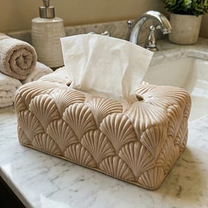 Op de afbeelding: Een beige tissuebox met een schelpenpatroon. Witte tissues steken uit de opening. De box staat op een badkamerblad, met andere badkameraccessoires op de achtergrond. De box is ongeveer 25 cm lang.