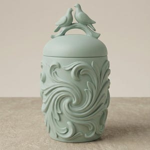 May include: A light green decorative jar with a lid, featuring a sculpted floral design. Two doves perch on a branch atop the lid. The jar has a cylindrical shape and is made of ceramic or a similar material.