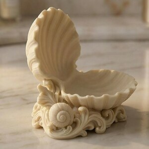 Puede incluir: Un plato decorativo en forma de concha de color crema. El plato est&aacute; abierto, revelando un peque&ntilde;o cuenco. La base presenta adornos y una concha. El plato est&aacute; hecho de un material liso, posiblemente cer&aacute;mico.
