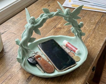 May include: A pale green decorative tray featuring two fairy figures supporting a vine arch. The tray contains a smartphone, keys, coins, and lip balm. It rests on a wooden table.