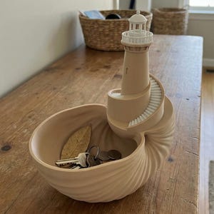 May include: A beige decorative lighthouse-shaped trinket dish. The lighthouse has a spiral staircase and sits atop a nautilus shell-shaped bowl. Keys and coins rest inside the bowl, with a dried leaf. The dish is on a wooden table.