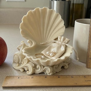 Puede incluir: Un joyero decorativo en forma de concha con una peque&ntilde;a bolsa con cord&oacute;n que contiene joyas, una manzana roja y una taza blanca. Una regla de madera se coloca junto a la taza. El joyero mide aproximadamente 15 cm de ancho.
