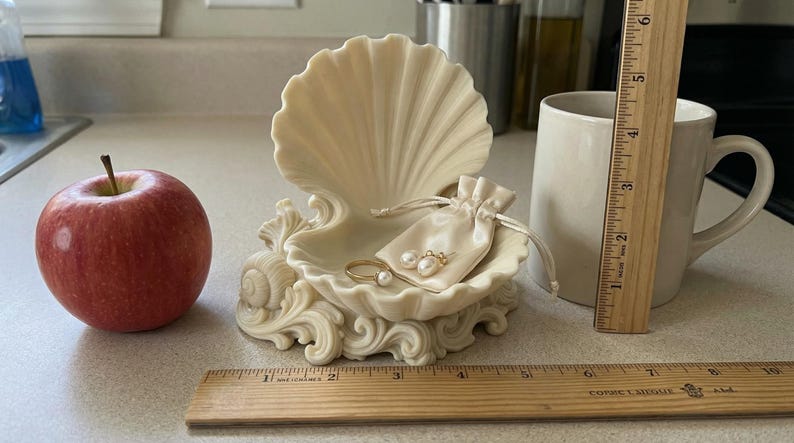 Puede incluir: Un joyero decorativo en forma de concha con una peque&ntilde;a bolsa con cord&oacute;n que contiene joyas, una manzana roja y una taza blanca. Una regla de madera se coloca junto a la taza. El joyero mide aproximadamente 15 cm de ancho.