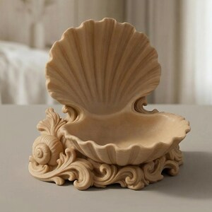 Puede incluir: Un plato decorativo beige con forma de concha. El plato presenta un dise&ntilde;o de concha grande y abierta con una superficie detallada y texturizada. La base est&aacute; adornada con volutas ornamentadas y un acento de concha, creando un tema costero.