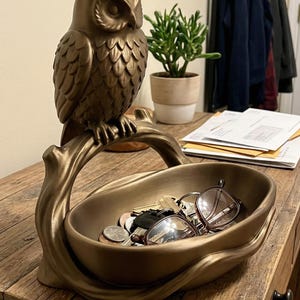 May include: A bronze-coloured owl-shaped catchall tray. The owl sits perched on a branch that forms the tray's handle. The tray holds keys, coins, and eyeglasses. The piece is on a wooden table.