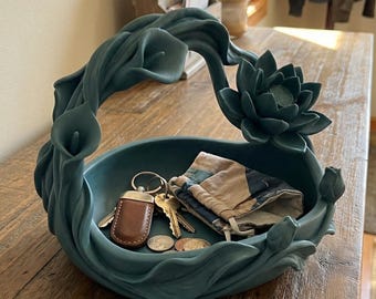 May include: A teal floral decorative bowl, holding keys, coins, and a small pouch, sits on a wooden table. The bowl's design incorporates calla lilies and a lotus flower. The bowl is a stylish storage solution.
