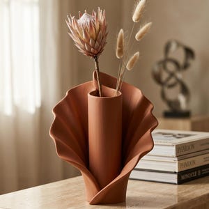May include: A terracotta-colored ceramic vase with a central cylindrical opening surrounded by wide, curved, fan-like petals. A single pink protea flower and three dried bunny tail grasses are arranged inside the central opening of the vase.