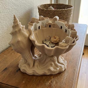 May include: A beige seashell-shaped decorative bowl with a miniature village scene inside. The bowl holds keys and a dried flower. The sculpture sits on a wooden table, showcasing a unique coastal or Mediterranean-inspired decor piece.