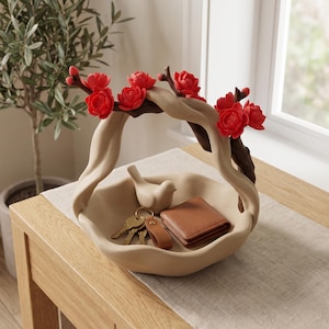 Op de afbeelding: Een beige keramieken kom met een golvende rand en een decoratief handvat in de vorm van een donkerbruine boomtak versierd met felrode bloemen. In de kom liggen een klein beige vogelbeeldje, een sleutelbos en een bruine lederen portemonnee.