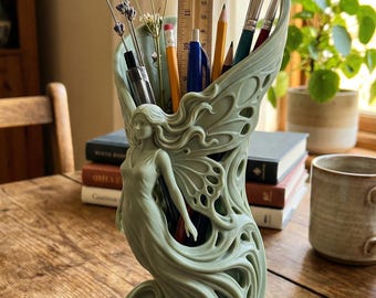 May include: A sage green fairy-themed desk tidy. The detailed sculpture holds pens, pencils, and brushes, with lavender and other flowers. The fairy has flowing hair and wings. A wooden ruler is visible. The organiser sits on a wooden table.
