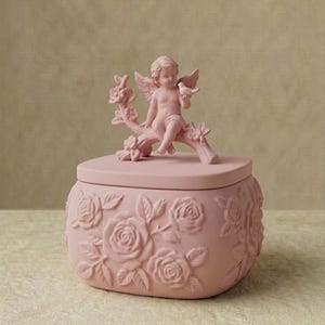 Könnte beinhalten: Rosa Keramik-Schmuckdose mit Deckel, mit einer geformten Engelsfigur, die einen Vogel auf einem Ast hält. Die Dose ist mit geprägten Rosenmotiven verziert. Ein dekorativer und romantischer Artikel.
