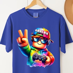 May include: Blue t-shirt featuring a colorful graphic of a character wearing a Roblox hat and sunglasses, holding a game controller. The character is making a peace sign. The word "ROBLOX" is printed on the hat.