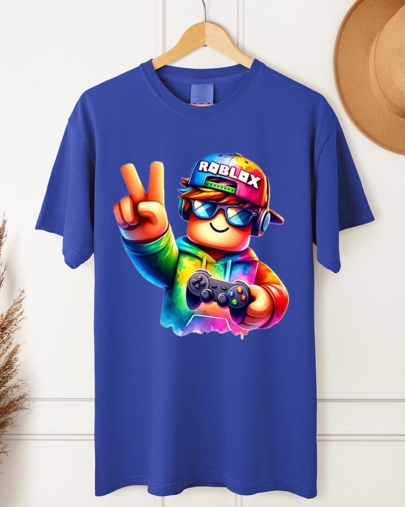 May include: Blue t-shirt featuring a colorful graphic of a character wearing a Roblox hat and sunglasses, holding a game controller. The character is making a peace sign. The word "ROBLOX" is printed on the hat.