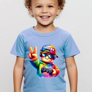 May include: A light blue t-shirt featuring a colorful graphic of a Roblox character wearing sunglasses and a hat, holding a game controller and making a peace sign. The word "ROBLOX" is on the hat.