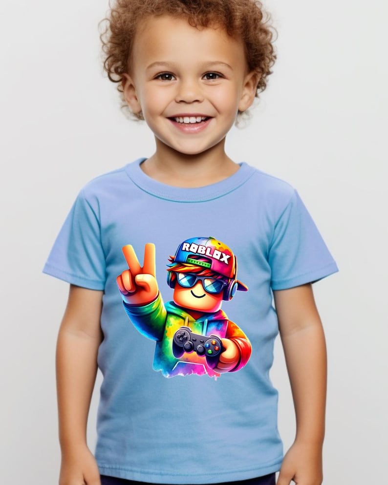 May include: A light blue t-shirt featuring a colorful graphic of a Roblox character wearing sunglasses and a hat, holding a game controller and making a peace sign. The word "ROBLOX" is on the hat.
