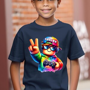 May include: Navy blue t-shirt featuring a colorful graphic of a Roblox character wearing a cap with the Roblox logo, sunglasses, and headphones, holding a game controller and making a peace sign.