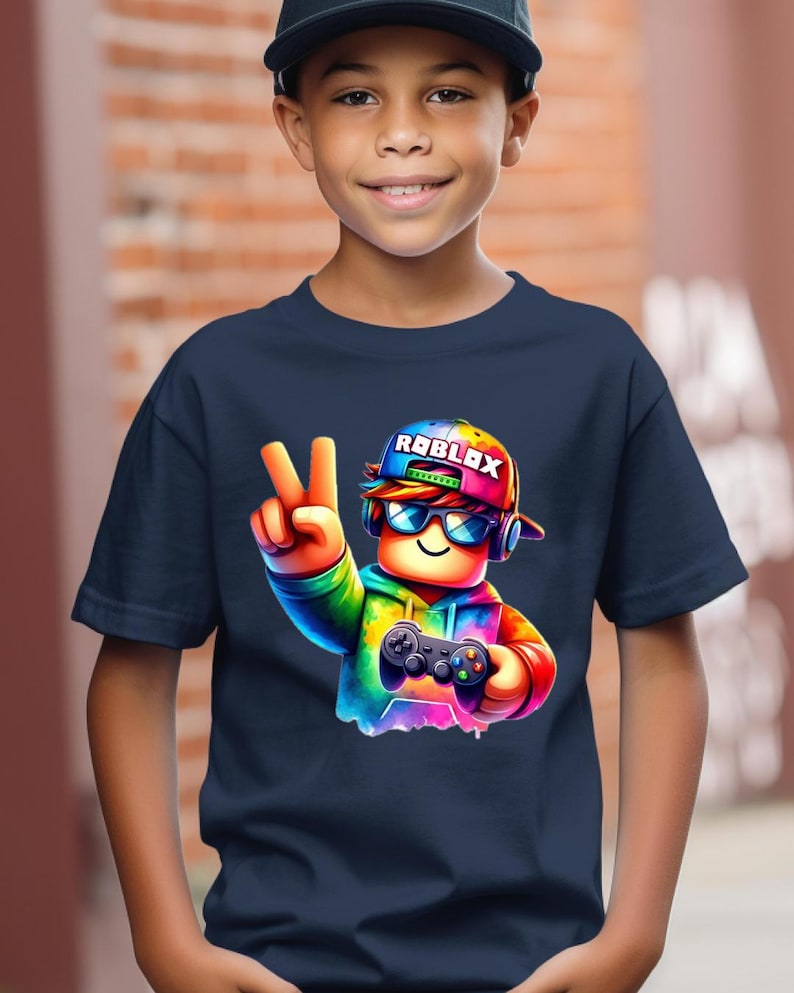May include: Navy blue t-shirt featuring a colorful graphic of a Roblox character wearing a cap with the Roblox logo, sunglasses, and headphones, holding a game controller and making a peace sign.