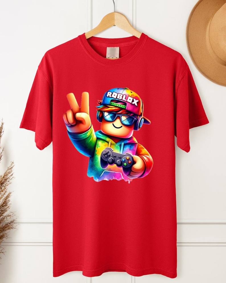 May include: Red t-shirt featuring a colorful cartoon character wearing a cap with the word "ROBLOX" and sunglasses, holding a game controller and making a peace sign. The character is rendered in vibrant rainbow hues.