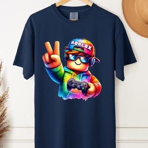May include: Navy blue t-shirt featuring a colorful cartoon character wearing a Roblox hat and sunglasses, holding a game controller and making a peace sign. The Roblox logo is visible on the hat. The design is vibrant and eye-catching.