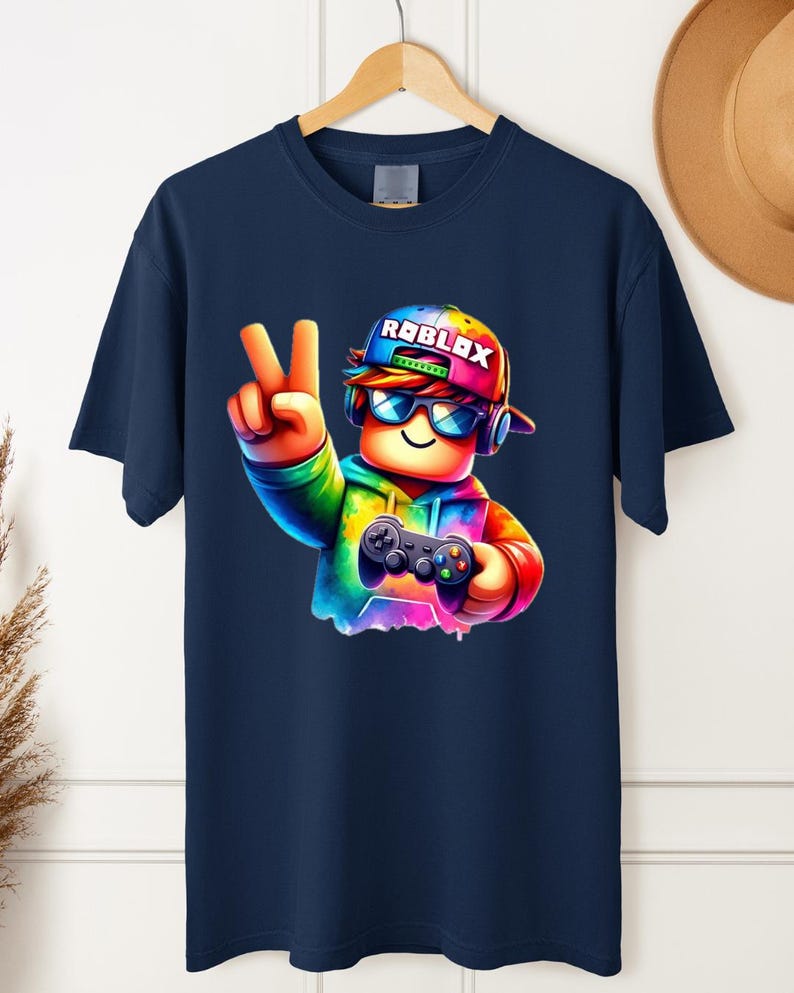May include: Navy blue t-shirt featuring a colorful cartoon character wearing a Roblox hat and sunglasses, holding a game controller and making a peace sign. The Roblox logo is visible on the hat. The design is vibrant and eye-catching.