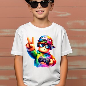 May include: White t-shirt featuring a colorful Roblox character wearing sunglasses, a cap with the Roblox logo, and holding a game controller. The character is making a peace sign with their fingers.