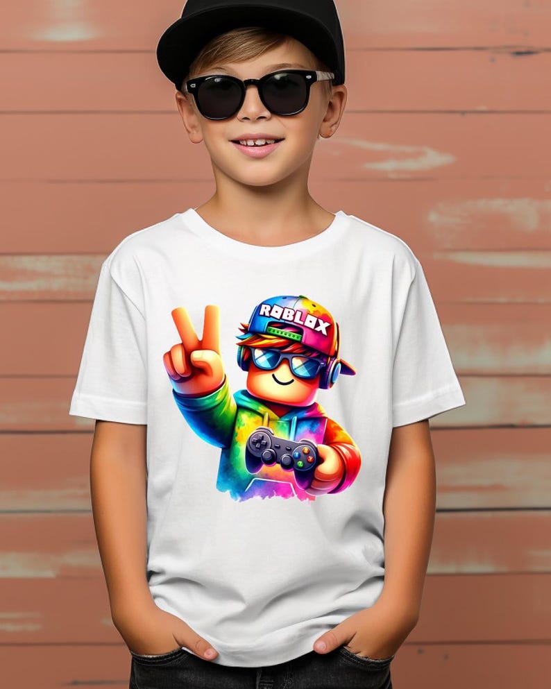 May include: White t-shirt featuring a colorful Roblox character wearing sunglasses, a cap with the Roblox logo, and holding a game controller. The character is making a peace sign with their fingers.