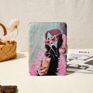 May include: A tablet case featuring a Scream mask with heart-shaped sunglasses and a pink boa, holding a drink with a heart-shaped straw and a pink telephone. The background has a soft, painted aesthetic.