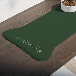 Könnte beinhalten: Eine dunkelgrüne Futtermatte für Haustiere in Knochenform mit dem Namen "Crosby" in weißer Schrift. Eine weiße Keramikschale mit Tierfutter steht auf der Matte. Die Matte liegt auf einem Holzboden neben einem gestreiften Teppich.