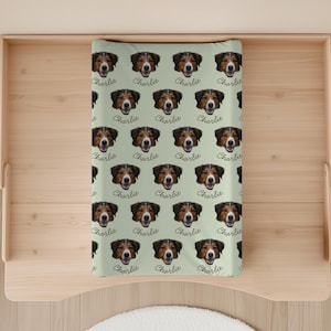 May include: A light green changing pad featuring a repeating pattern of a dog's face with the name "Charlie" in a cursive font. The dog has brown and white fur.