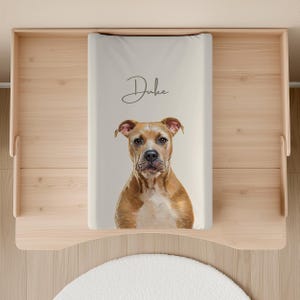 Peut inclure: Un matelas à langer beige clair avec le nom "Duke" en écriture cursive. Le matelas présente un portrait d'un chien brun et blanc. Le matelas est posé sur une table à langer en bois clair.