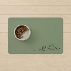 Puede incluir: Un tapete verde salvia para comida de mascotas con un cuenco moteado blanco y gris lleno de croquetas marrones. El tapete tiene el nombre "Bella" en una fuente de escritura negra.