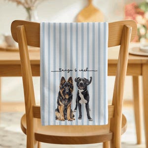 May include: A waffle-weave kitchen towel featuring vertical light blue and white stripes. The towel displays a printed illustration of a German Shepherd and a black dog with a white chest, with the text "tango & cash" written in black script.