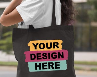 May include: Black tote bag with the text "YOUR DESIGN HERE" in white letters over three horizontal brush strokes of yellow, magenta, and teal. The bag is carried over a person's shoulder.