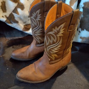 Puede incluir: Un par de botas vaqueras marrones a media pantorrilla con punta afilada, tacón y tiradores en la parte superior. La caña de cada bota está decorada con intrincados patrones de bordado de color claro y tiene un forro interior de color naranja brillante.