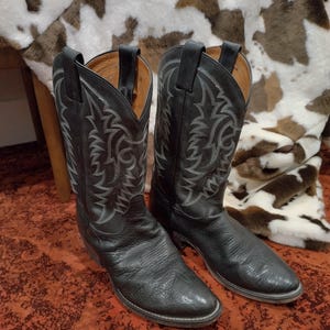 Op de afbeelding: Een paar zwarte leren cowboylaarzen met een puntige neus, een blokhak en ingewikkelde grijze decoratieve stiksels op de hoge schacht. Elke laars is voorzien van twee treklipjes bij de opening aan de bovenkant en een textuur op het leer.