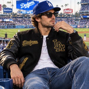 Könnte beinhalten: Eine königsblaue Baseballkappe mit weißem LA-Logo, eine schwarze Sonnenbrille und eine schwarze Collegejacke mit gelber Dodgers- und LA-Stickerei. Die Jacke hat schwarze Lederärmel, ein weißes T-Shirt darunter und eine blaue Jeans.