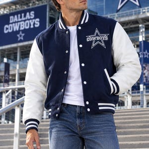 Pode incluir: Uma jaqueta varsity azul-marinho e branca com o logotipo da estrela do Dallas Cowboys no peito, mangas brancas, fecho frontal com botões de pressão e acabamento canelado listrado na gola, punhos e bainha, sobre uma camiseta branca e jeans.
