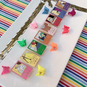 May include: A collection of colorful butterfly hair clips and illustrated cards arranged on a white tray. The cards feature various illustrations and text, with a pink, yellow, green, orange, and purple color scheme. The background includes a striped fabric.